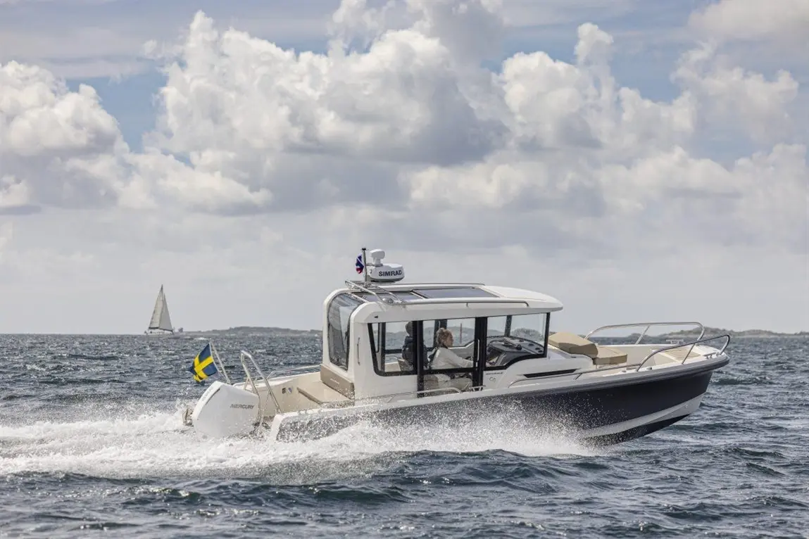A motorboat with a Swedish flag speeds through waves, with a sailboat in the distance and a cloudy sky above.